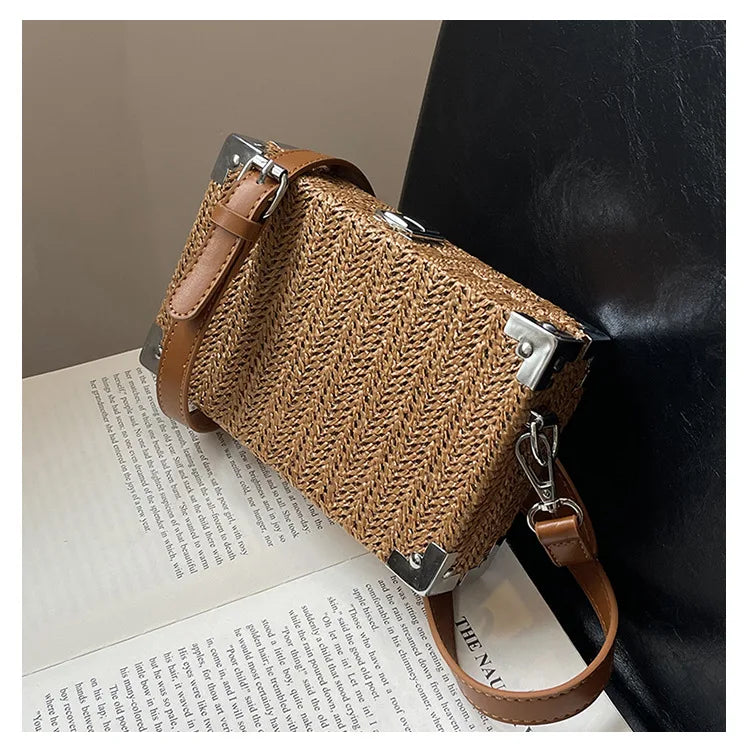 Borsa a tracolla in paglia intrecciata con angoli in metallo e cinturino in pelle marrone, posata su un libro aperto. Elegante e alla moda.