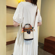 Donna con abito bianco indossa borsa a secchiello beige con dettagli colorati e tracolla marrone. Accessorio moda elegante e trendy.