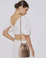 Donna con borsa secchiello beige, scritta "The Bucket", indossa abito bianco elegante. Accessorio moda femminile, stile chic e moderno.