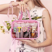 Borsa trasparente rosa con scritta "STUFF", contenente cosmetici e accessori, tenuta da una donna in abito floreale. Accessori moda, organizer trucco.