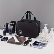 Borsa da toilette nera con manici, circondata da prodotti per la cura personale e cosmetici su sfondo grigio. Accessori da viaggio eleganti.