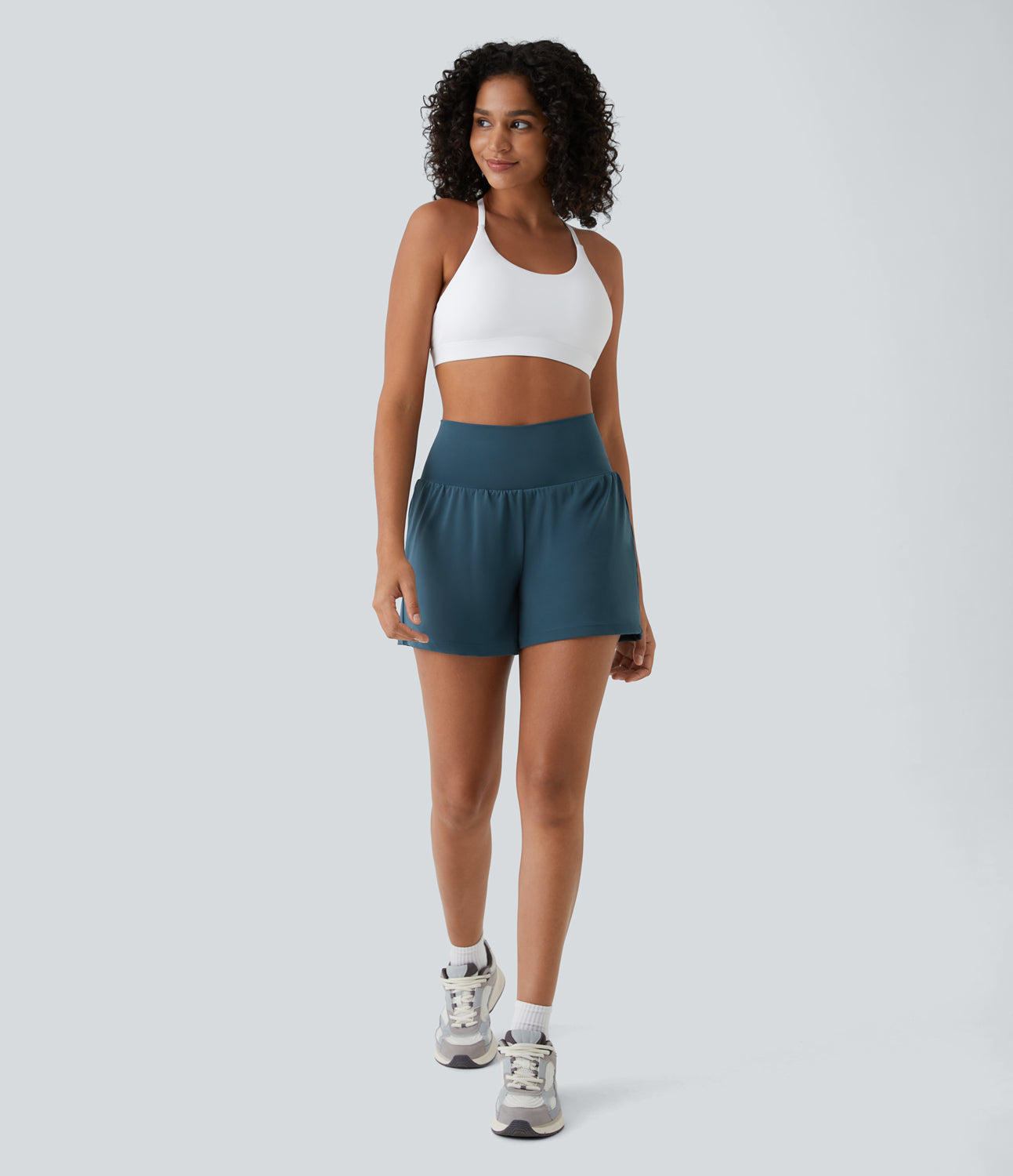 Donna in abbigliamento sportivo: top bianco, pantaloncini blu, scarpe da ginnastica grigie. Abbigliamento fitness, moda sportiva donna.