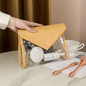 Borsa trasparente in PVC con dettagli in pelle gialla, contenente cosmetici e accessori da trucco, su tavolo con tazza e pennelli da trucco.