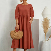 Abito lungo rosso terracotta con maniche a tre quarti, borsa in paglia intrecciata, sfondo bianco minimalista, moda donna elegante.