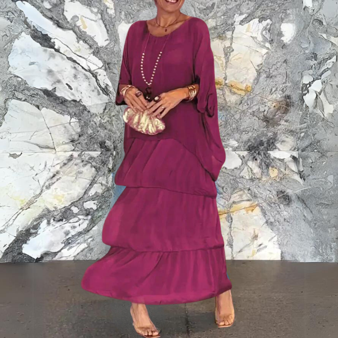 Abito lungo viola a balze, indossato da una donna con borsa a mano e collana, su sfondo marmo. Moda elegante, abbigliamento femminile chic.