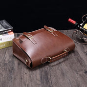 Borsa a tracolla in pelle marrone su tavolo di legno, con fibbie dorate. Elegante accessorio moda uomo, ideale per lavoro e viaggi.
