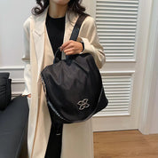 Donna con cappotto beige tiene borsa nera elegante con logo distintivo, ideale per moda autunno-inverno. Accessorio chic per look sofisticato.