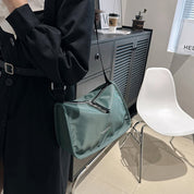 Borsa a tracolla verde in nylon, design moderno, indossata da una persona in abito nero. Sfondo con sedia bianca e mobile grigio.