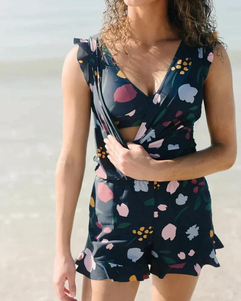 Donna in spiaggia con tuta estiva floreale blu scuro, scollo a V, senza maniche. Moda mare, abbigliamento estivo donna, look casual elegante.