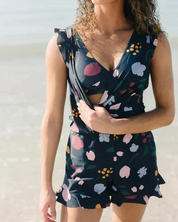 Donna in spiaggia con tuta estiva floreale blu scuro, scollo a V, senza maniche. Moda mare, abbigliamento estivo donna, look casual elegante.