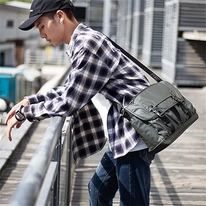 Giovane con camicia a quadri e cappello nero indossa borsa a tracolla verde militare, appoggiato a una ringhiera in ambiente urbano.