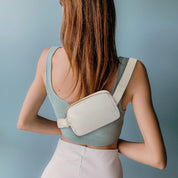 Donna con marsupio bianco, indossa top verde e gonna bianca. Moda casual, accessorio trendy, stile estivo. Sfondo blu chiaro.