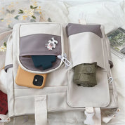 Zaino beige con tasche multiple, smartphone giallo, portachiavi mucca, ombrello pieghevole verde, su letto bianco. Accessori moda, organizzazione viaggio.