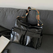 Borsa a tracolla nera in pelle lucida su divano grigio, con tasche frontali e tracolla decorata. Accessorio moda elegante e pratico.