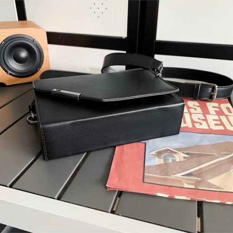 Borsa a tracolla in pelle nera su tavolo con altoparlante in legno e rivista. Elegante accessorio moda uomo, design moderno e funzionale.