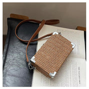 Borsa a tracolla in rattan intrecciato su sedia nera, design rettangolare, dettagli in metallo argentato, stile boho chic, moda donna.