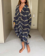 Donna che indossa un abito lungo a righe tie-dye blu e gialle, con bottoni e maniche lunghe, in piedi su un pavimento chiaro. Moda casual elegante.