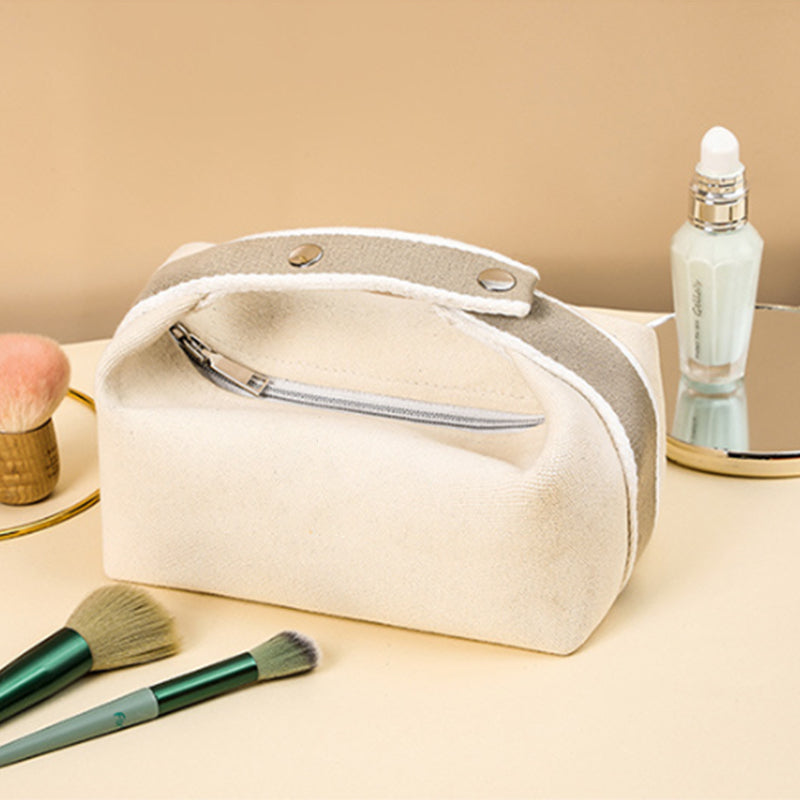 Borsa cosmetica in tela beige con cerniera, circondata da pennelli trucco e flacone di siero su sfondo neutro. Accessorio da viaggio elegante.