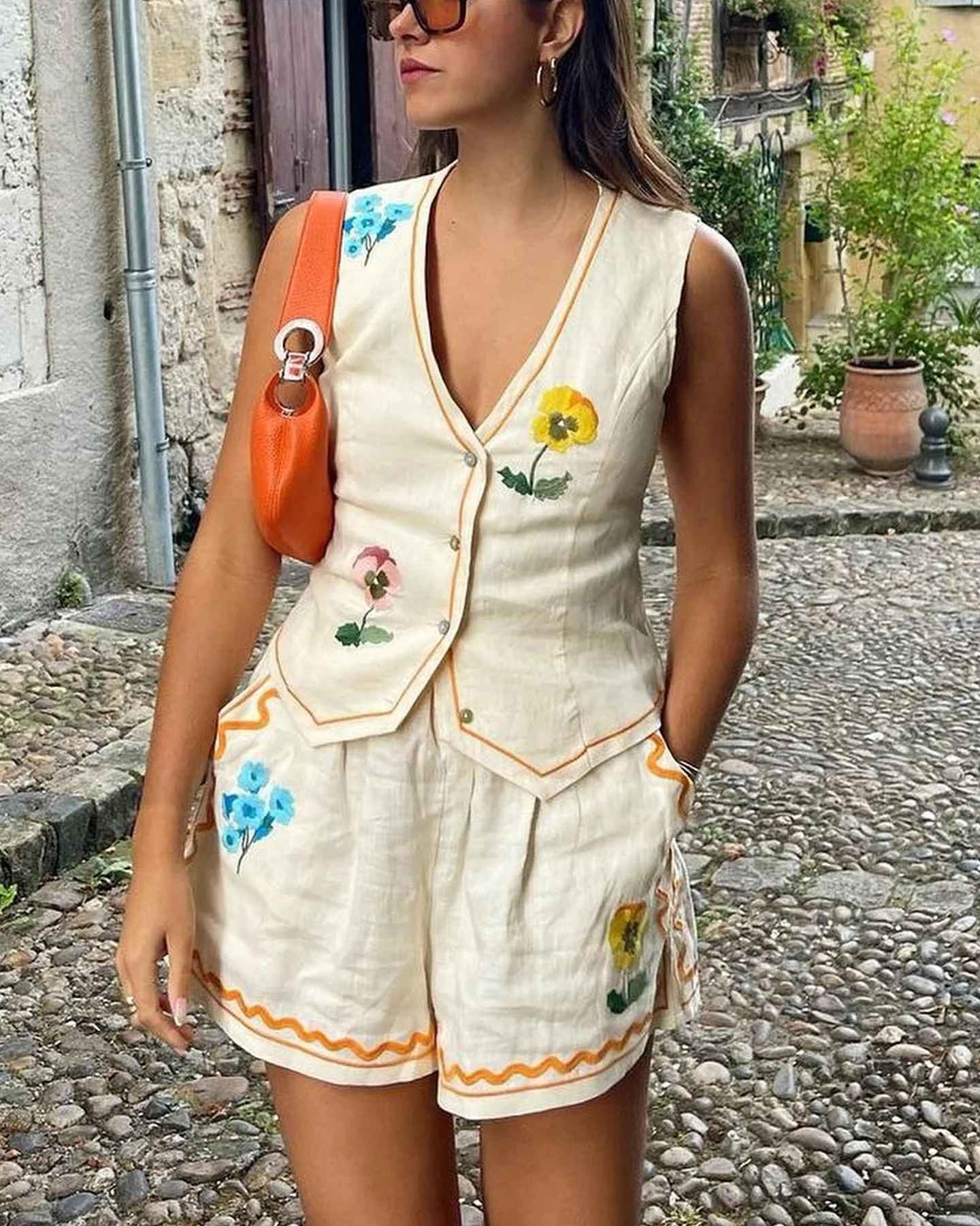 Donna con completo estivo in lino bianco con fiori colorati, borsa arancione, occhiali da sole, su strada acciottolata. Moda estiva casual elegante.