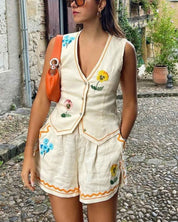 Donna con completo estivo in lino bianco con fiori colorati, borsa arancione, occhiali da sole, su strada acciottolata. Moda estiva casual elegante.