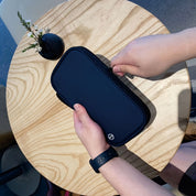 Mani che aprono un astuccio nero su un tavolo di legno chiaro, accanto a un piccolo vaso con fiore. Accessorio elegante, pratico per organizzazione.