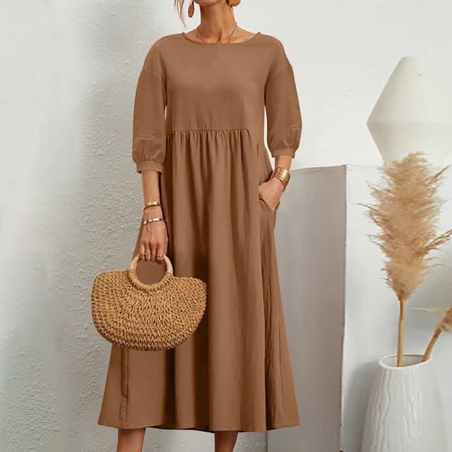 Abito lungo marrone con maniche a tre quarti, borsa in paglia intrecciata, accessori dorati, sfondo bianco minimalista, pianta decorativa.