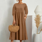 Donna con abito marrone lungo, maniche a tre quarti, borsa in paglia intrecciata, sfondo bianco minimalista, decorazioni naturali.