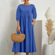 Donna con abito blu a maniche lunghe, borsa di paglia intrecciata, sfondo bianco minimalista, decorazione pampas. Moda estiva elegante.