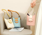 Tre borse a secchiello in pelle color crema, azzurro e rosa con fibbia decorativa, su sedia beige. Accessori moda eleganti per donna.