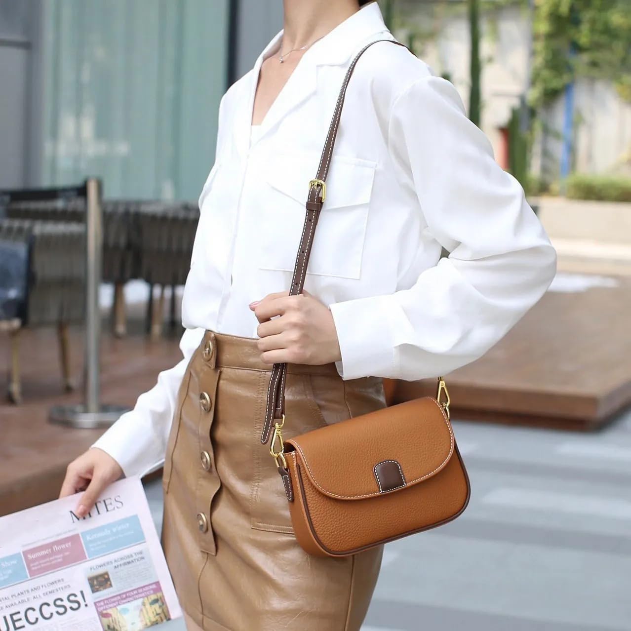 Donna con camicia bianca e gonna marrone, tiene una borsa a tracolla in pelle marrone chiaro. Accessorio moda elegante e casual.