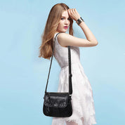 Donna con abito bianco indossa borsa a tracolla nera in pelle, sfondo azzurro. Elegante accessorio moda femminile, stile casual chic.