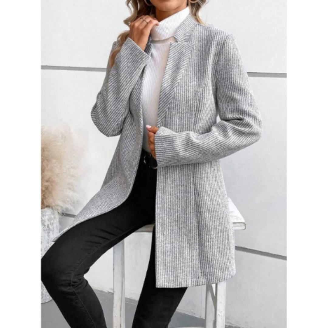 Giacca lunga grigia a righe verticali, indossata su maglione bianco a collo alto, abbinata a pantaloni neri. Look elegante e moderno per donna.