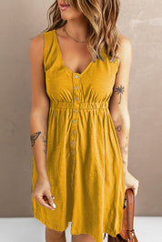 Donna con abito giallo senape senza maniche, bottoni frontali, tatuaggi visibili su braccia, sfondo neutro. Moda estiva casual elegante.