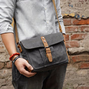 Borsa a tracolla in tela grigia con chiusura in pelle marrone, indossata da uomo in camicia grigia. Accessorio moda casual, ideale per uso quotidiano.