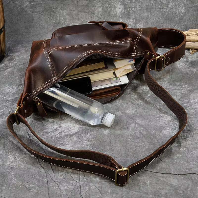 Borsa a tracolla in pelle marrone aperta, contenente bottiglia d'acqua, libri e smartphone. Design elegante, ideale per lavoro e viaggio.