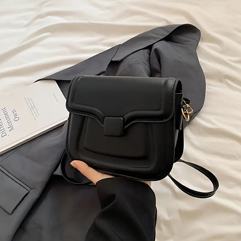 Borsa a tracolla nera in pelle su letto bianco, accanto a giacca grigia e libro. Design elegante, ideale per moda donna e accessori di tendenza.