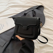 Borsa a tracolla nera in pelle su letto bianco, accanto a giacca grigia e libro. Design elegante, ideale per moda donna e accessori di tendenza.