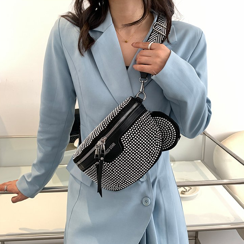 Donna con blazer azzurro indossa borsa a tracolla nera con borchie bianche, cerniera e tracolla decorata. Accessorio moda elegante e moderno.
