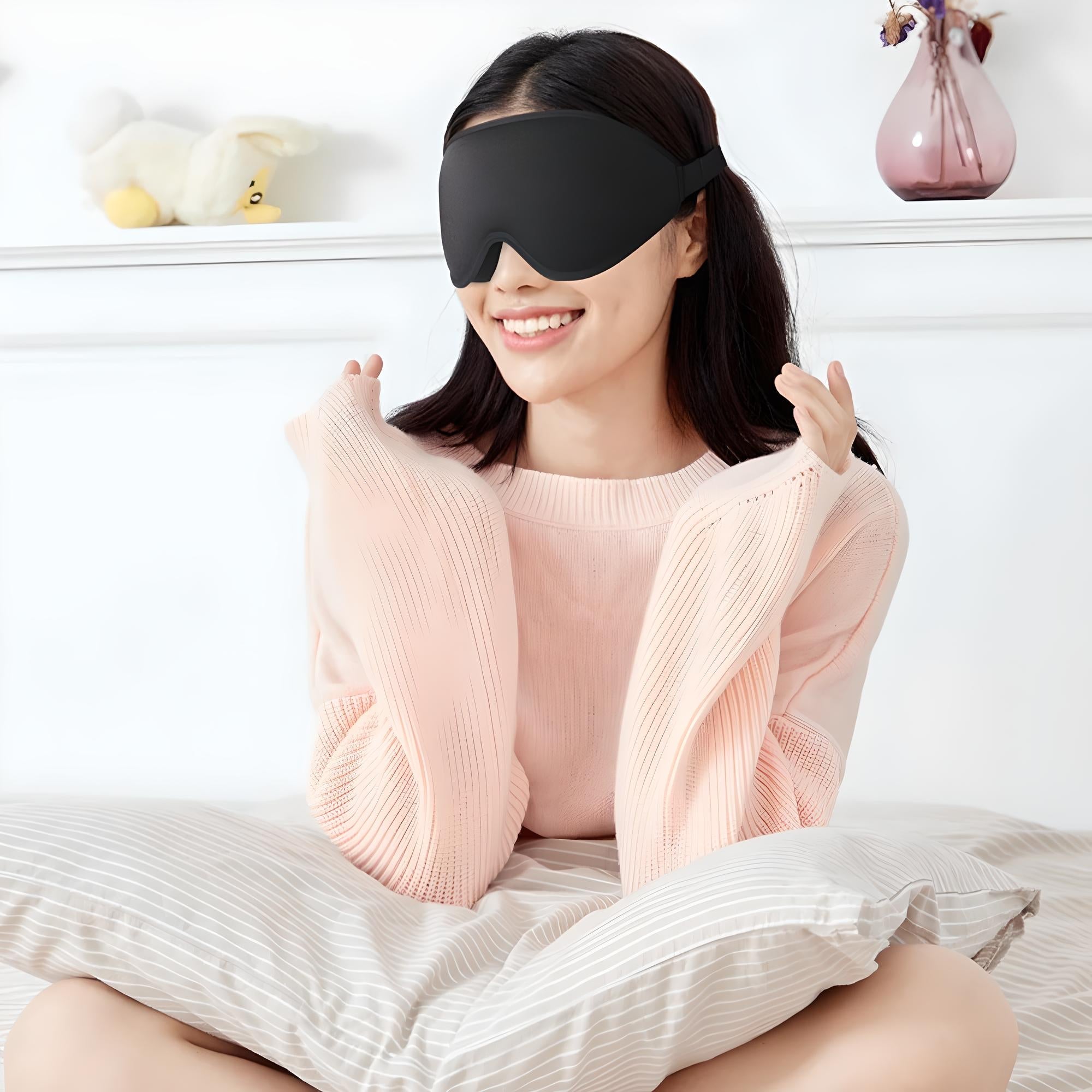 Donna sorridente con maschera per dormire nera, seduta su letto con cuscino a righe, indossa maglione rosa. Relax, comfort, accessori notte.
