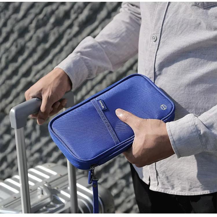 Uomo con camicia grigia tiene un portafoglio da viaggio blu accanto a una valigia argentata. Accessorio organizzatore per documenti e passaporti.