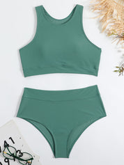 Costume da bagno verde a due pezzi, top sportivo e slip a vita alta, su sfondo bianco con occhiali e accessori estivi.