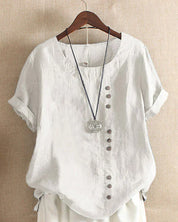 Camicia bianca in lino da donna con bottoni decorativi asimmetrici, maniche corte risvoltabili, su gruccia di legno. Stile casual ed elegante.