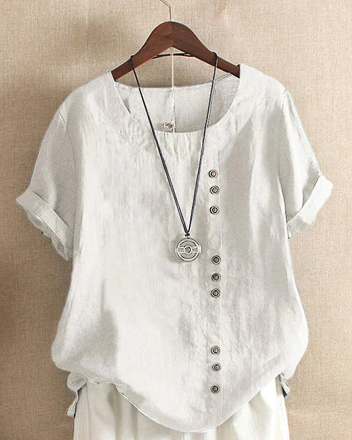 Camicia bianca in lino da donna con bottoni decorativi asimmetrici, maniche corte risvoltabili, su gruccia di legno. Stile casual ed elegante.