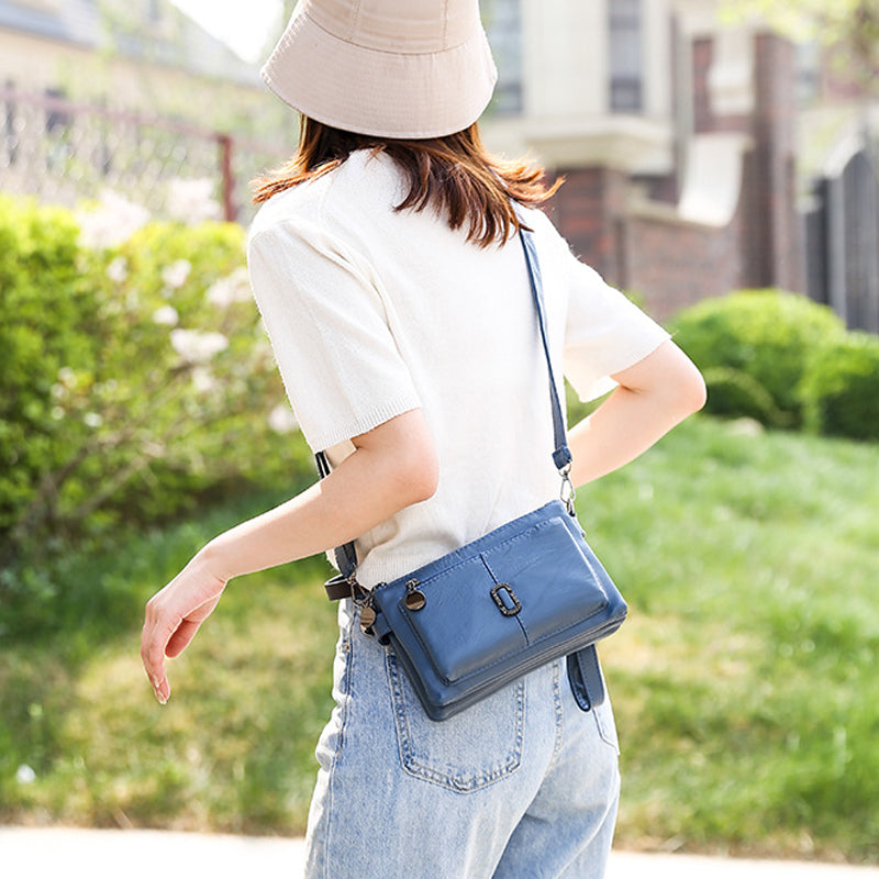 Donna con borsa a tracolla blu in pelle, indossa cappello beige, maglietta bianca e jeans chiari, passeggia in un giardino soleggiato.