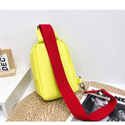 Zaino giallo con tracolla rossa su tavolo bianco, accanto a cornice e rivista. Design moderno, ideale per moda urbana e casual.