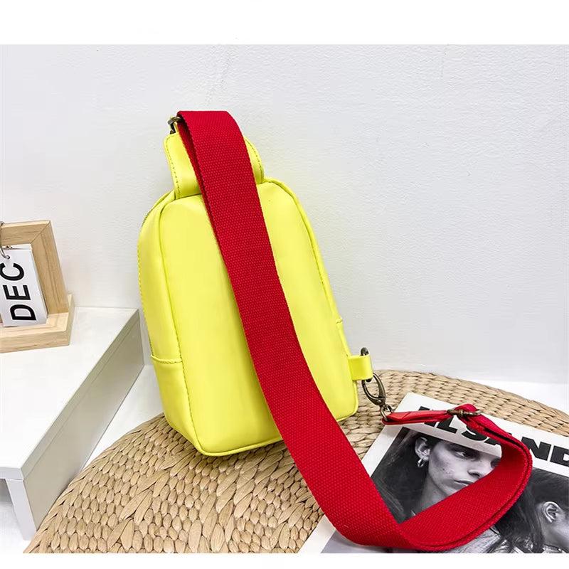 Zaino giallo con tracolla rossa su tavolo bianco, accanto a cornice e rivista. Design moderno, ideale per moda urbana e casual.
