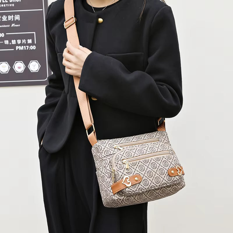 Borsa a tracolla beige con motivo geometrico, dettagli in pelle marrone e cerniere dorate, indossata da una persona in cappotto nero.