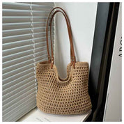 Borsa a tracolla in paglia intrecciata beige con manici in pelle marrone, appoggiata su una sedia vicino a una finestra con persiane grigie.