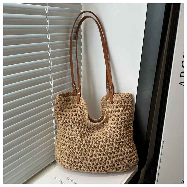 Borsa a tracolla in paglia intrecciata beige con manici in pelle marrone, appoggiata su una sedia vicino a una finestra con persiane grigie.
