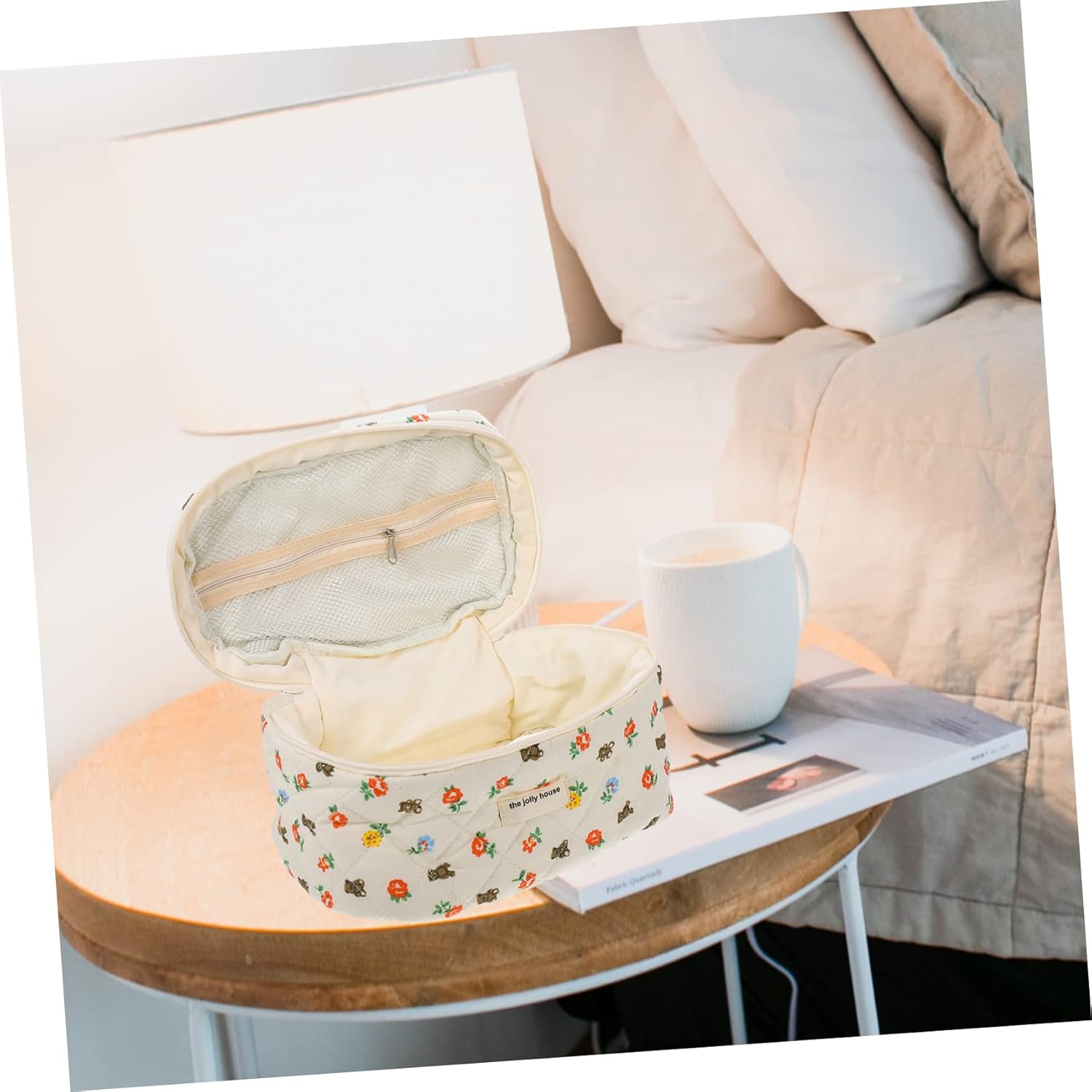 Astuccio da viaggio floreale aperto su tavolino accanto a letto, con lampada e tazza bianca. Design elegante, ideale per cosmetici e accessori.